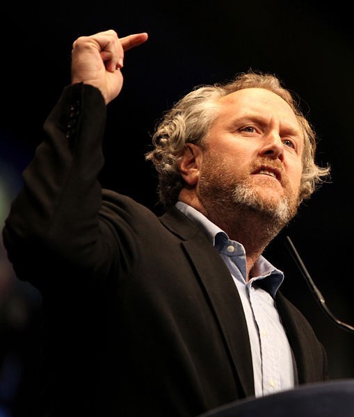Photo of Andrew Breitbart, in a dark suit and graying beard, speaking with an arm in the air