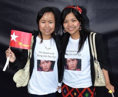 Two women smile as they wave a flag and wear shirts with Aung San Suu Kyi's photo on them