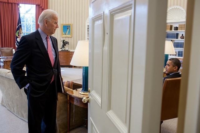 Photo of the oval office, with Joe Biden in a blue suit talking to Barack Obama, viewed through the open door