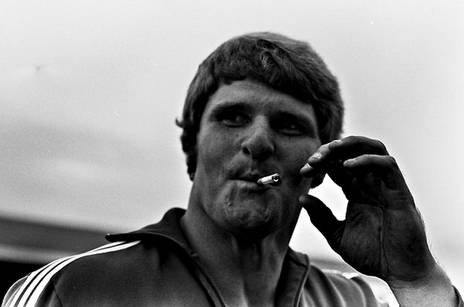 A big strong man in a track suit smokes a Marlboro cigarette, grinning