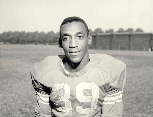 A very young Bill Cosby wears an old-fashioned football jersey, number 39, as he stands on a practice field