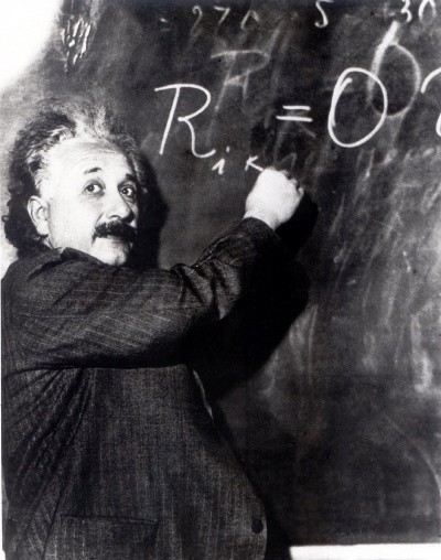 A photo of Albert Einstein at a chalkboard, writing an equation and looking back at the room