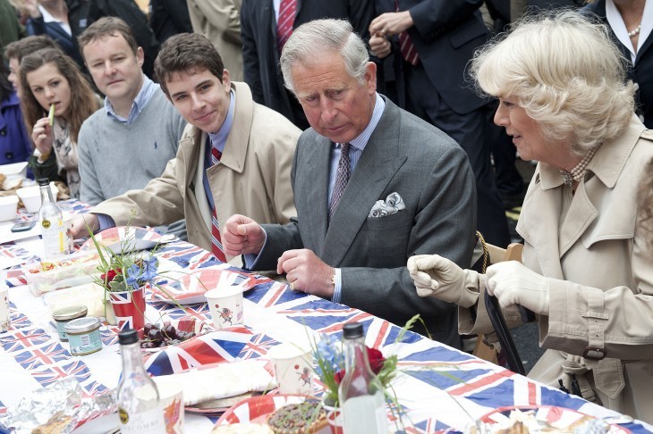 Photo of Prince Charles and Camilla sitting at a long lunch table with Union Jack tablecloth, chatting with commoners