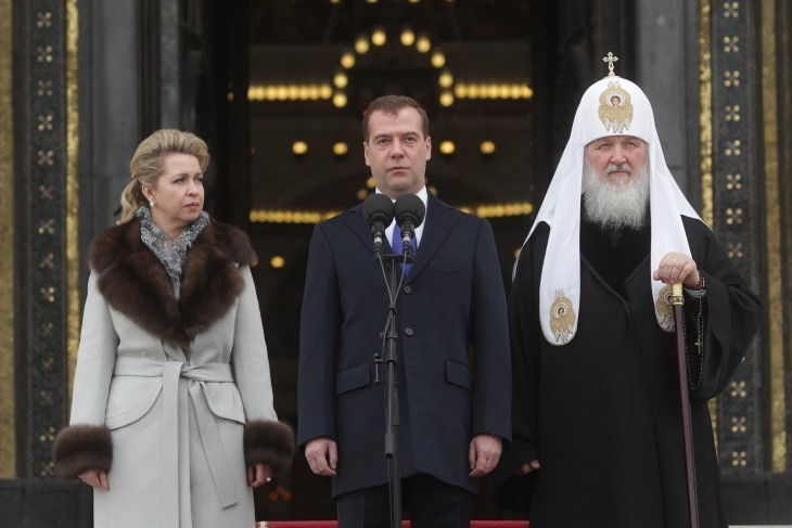 Photo of the Patriarch of Moscow, in a fancy white hat, standing next to a politician in a blue suit and his wife
