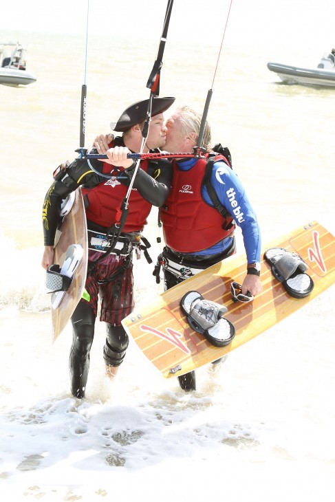 Photo of Richard Branson kissing his son, Sam, as they both stand in wetsuits in the surf with kite surfing gear