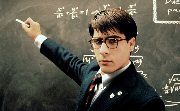 A photo of Jason Schwartzman in a prep school blazer and funny glasses, writing equations on a chalkboard