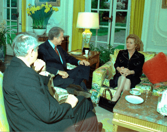 Photo of Margaret Thatcher on a flowery couch in a sitting room, chatting with Jimmy Carter and Cyrus Vance