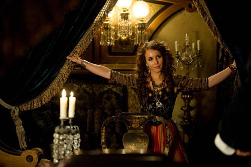 Photo of Noomi Rapace as a gypsy-type woman, holding open curtains and smiling alluringly