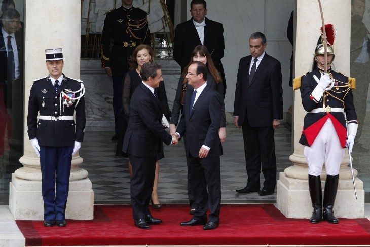 Nicolas Sarkozy and Francois Hollande, in business suits, shake hands between formal guards on a red carpeted stairway