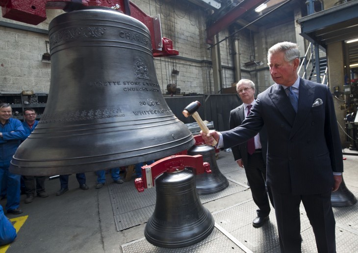 Photo of Prince Charles, using a medium-sized mallet to whack a hanging bronze bell the size of, say, a dishwasher