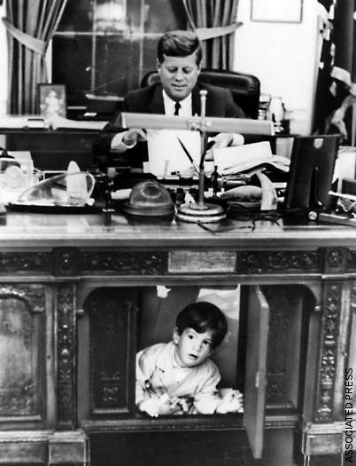 A young boy pops through a panel in the leg hole of JFK's big desk