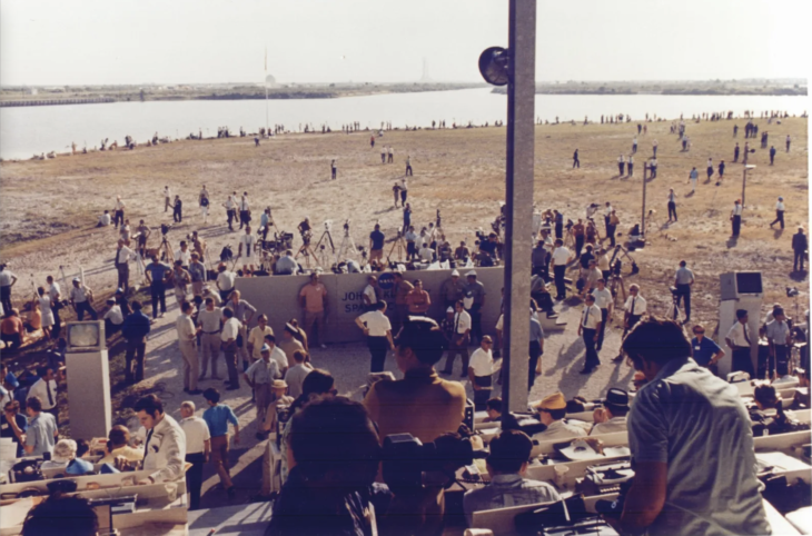 Crowds mill about on the edge of water with a rocket in the distance