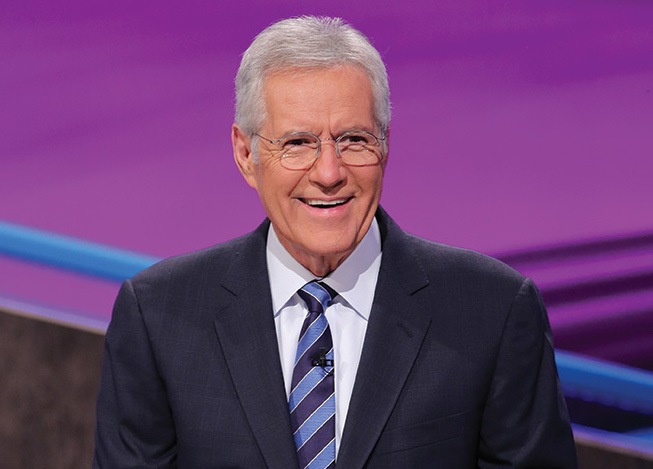 Alex Trebek wears his classic business suit and tie; his hair is gray. He is smiling broadly as he looks a bit to his left.