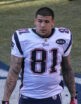 Aaron Hernandez walks across a football field. He is muscular, with short hair; both arms are covered with tattoos, and he wears red receiver's gloves.