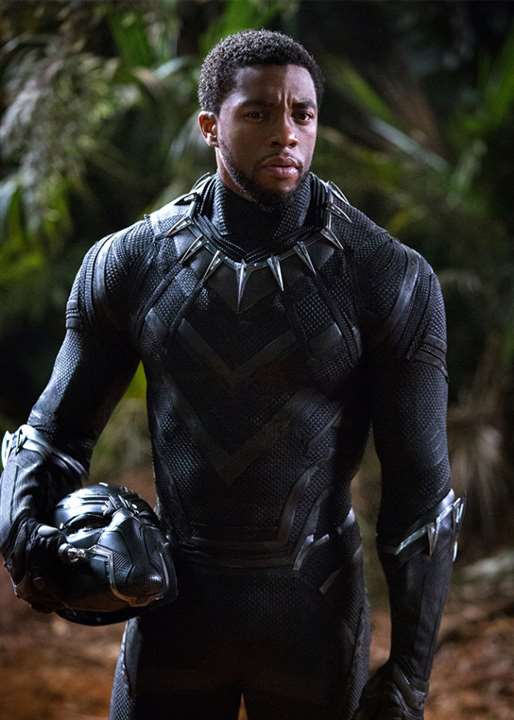 Chadwick Boseman stands in a jungle, wearing a sleek black superhero suit and holding a mask/helmet in his hand. He looks terrific and heroic.