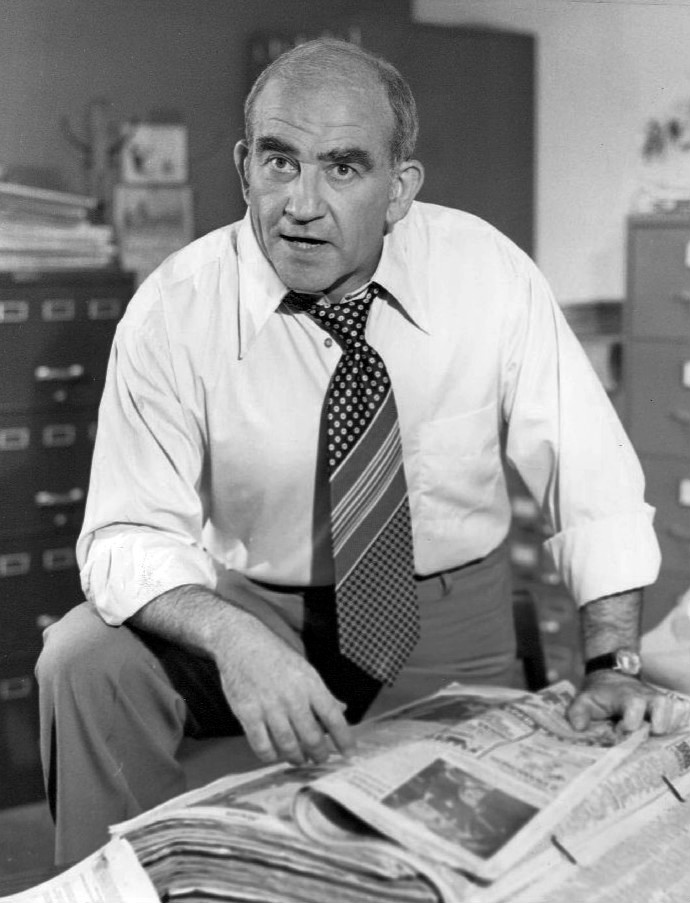 Ed Asner is a balding man with a wide 1970s tie and a long-sleeved white business shirt; he stands in front of newspaper clippings in a newsroom, where he appears to be barking orders to someone