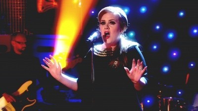 Photo of Adele onstage, with colored lights, holding her hands up as she sings
