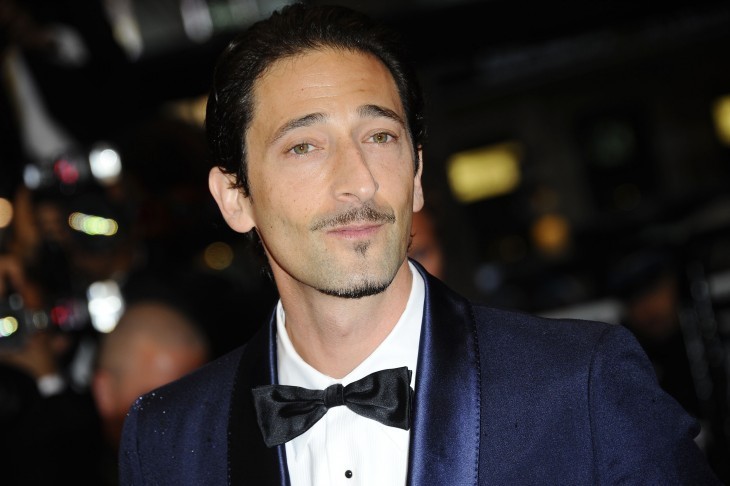 Adrien Brody in a blue tuxedo and black tie, tilting his head at the camera, lips closed