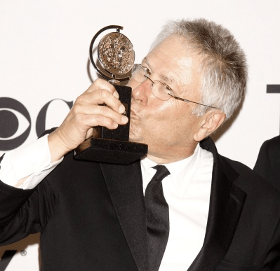 A photo of Alan Menken kissing his Tony Award gleefully backstage, in a formal black suit (Menken, not the Tony)