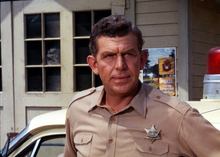 A photo of Andy Griffith in his sheriff's uniform in Mayberry, looking left as he stands by a police car