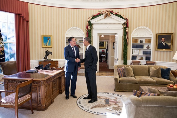 Photo of Barack Obama shaking Mitt Romney's hand, next to the president's desk in the oval office, both men wearing blue suits