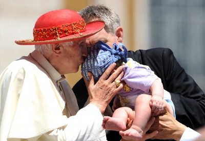 Photo of Pope Benedict XVI, in red hat, kissing an infant in a bonnet in the sunshine