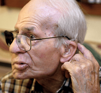 A photo of an elderly man with a hearing aid and gray hair, pointing to the back of his head