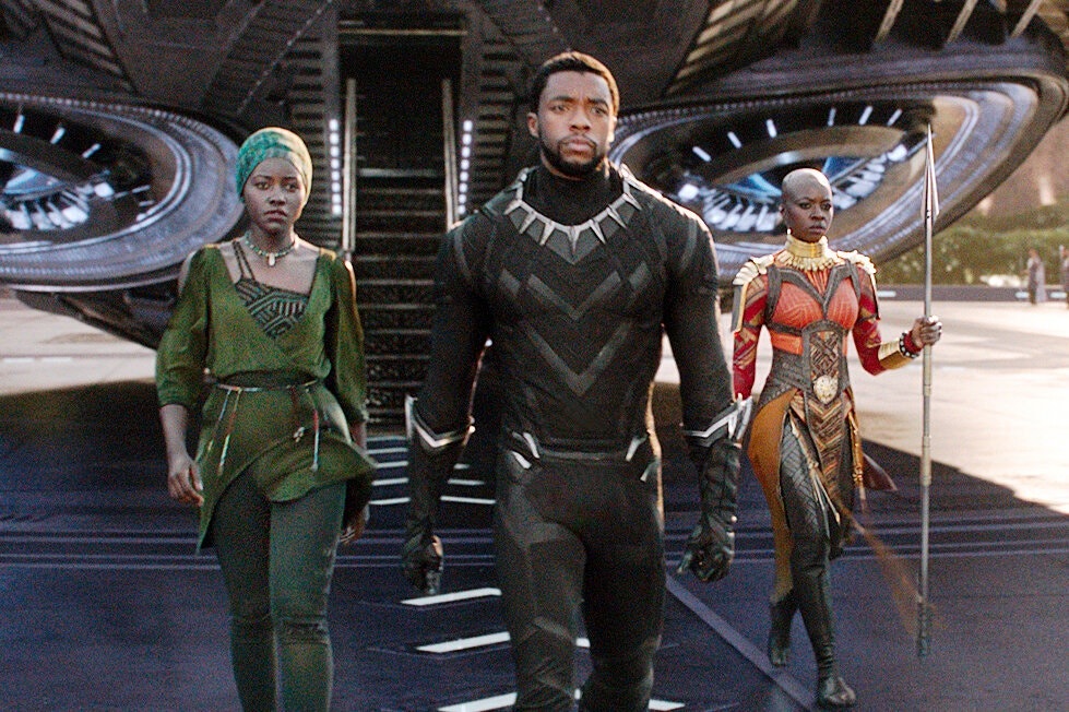 Chadwick Boseman wears a sleek, skin-tight black suit and is flanked by two Black women in exotic African dress. They seem to have just descended the stairs of a spaceship or super-plane of some kind