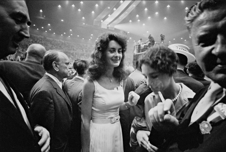 A photo of a woman in a white dress surrounded by smoke and bustle on the convention floor