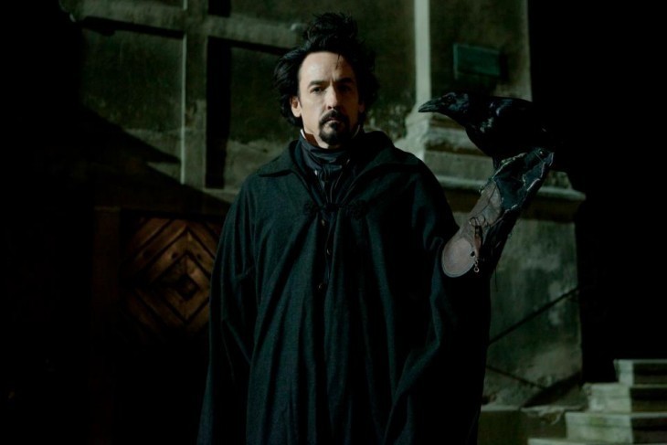A photo of John Cusack holding a raven on his arm, dressed in 19th-century finery (Cusack, not the raven)