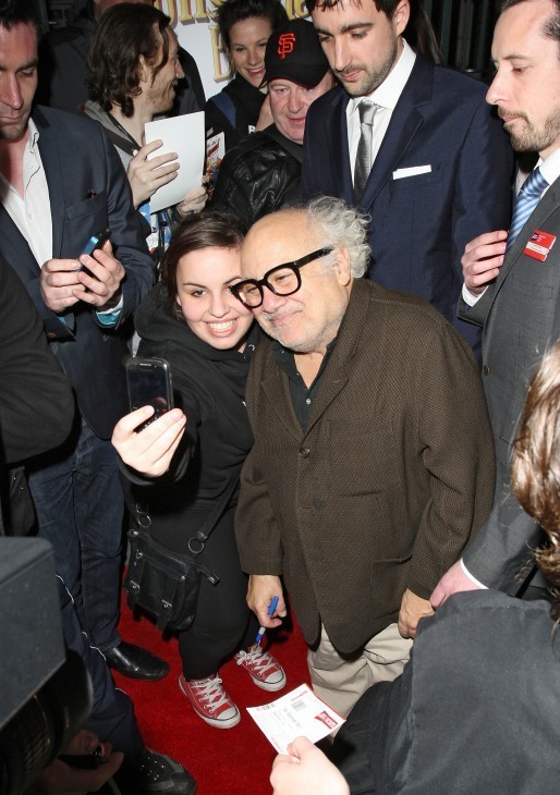 A photo of Danny DeVito, white-haired and grinning, leaning in with a fan who holds a cell phone camera at arm's length
