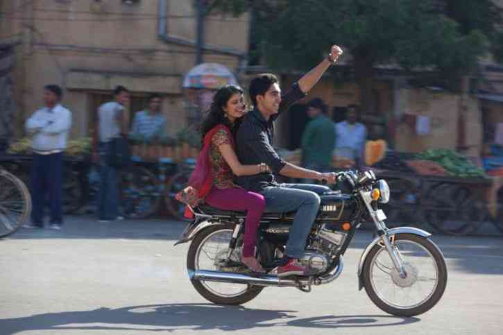 Photo of Dev Patel riding a motorcycle with his smiling girlfriend clinging to the back. In India!
