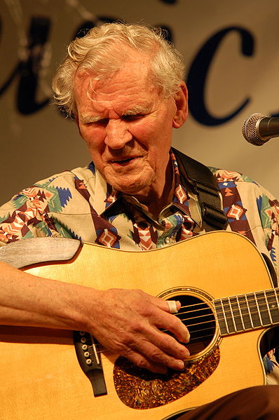 Photo of Doc Watson strumming a guitar and singing onstage in a handsomely gaudy shirt