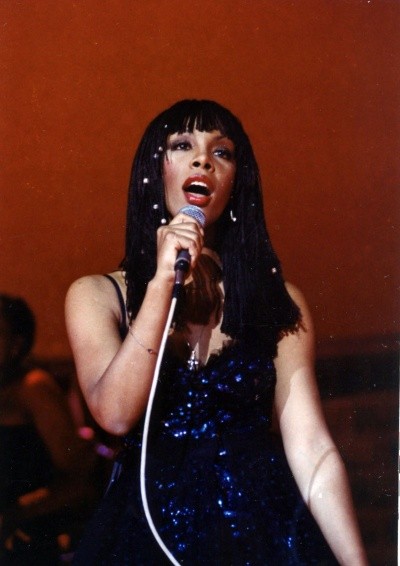 A photo of Donna Summer in a sparkly dress, holding up a microphone as she sings onstage