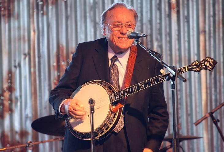 A photo of Earl Scruggs, with a banjo, smiling at the crowd in front of a corrugated tin backdop