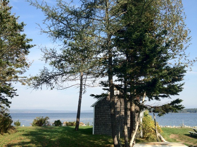 A small shack with cedar shingles sits on the shore of a wide bay