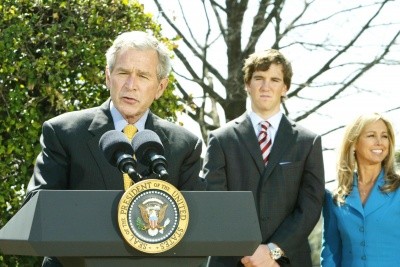 President Bush speaks at an outdoor podium while Eli Manning listens behind him