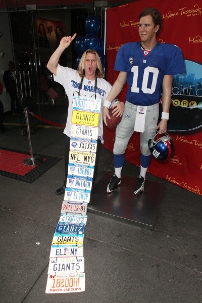 A nutty-looking man holding a long string of license plates stands next to a wax statue of Eli Manning