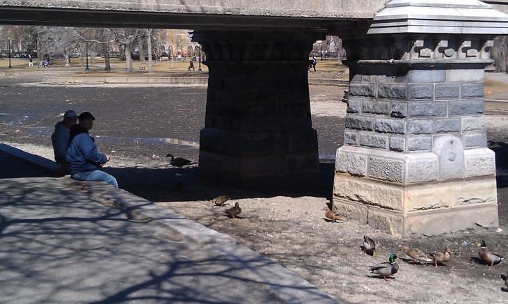 Two men sit under the bridge and toss bread to ducks on the muddy lake bottom