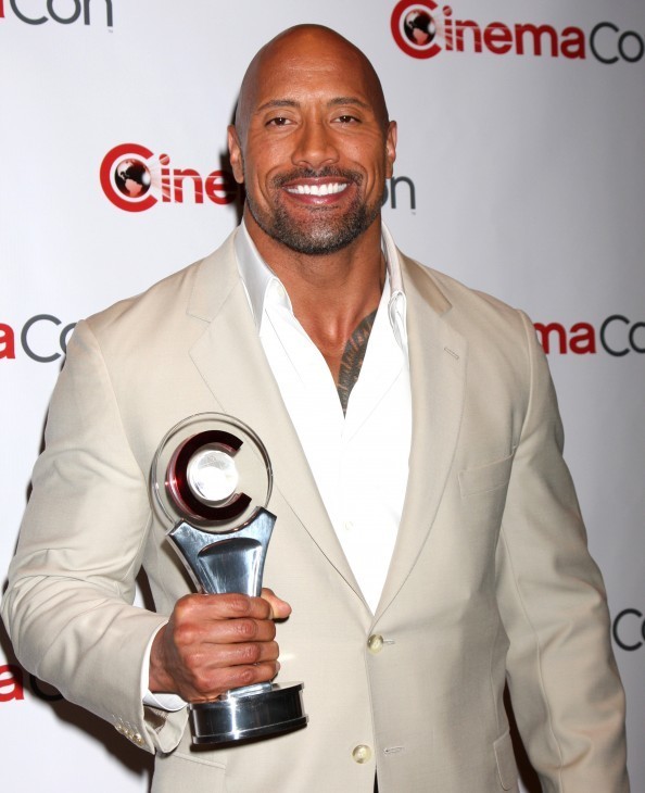 Photo of The Rock, gripping a movie-type award trophy, and smiling for cameras in a white suit