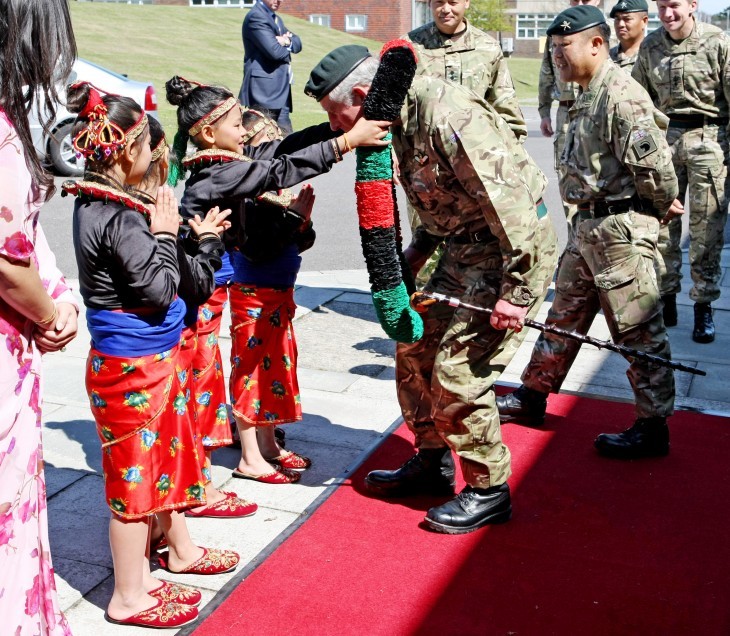 Photo of Prince Charles in military fatigues, bending over as a child puts a huge wreath or lei over his head