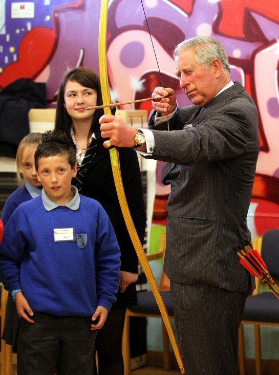 Prince Charles draws back on a bow and arrow, peering closely, as children smile and look on