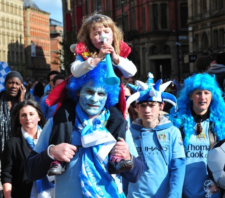 A silly and funny photo of a family with faces painted blue (for Manchester City) and wearing funny hats at the parade