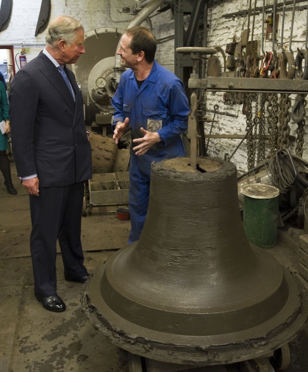 Photo of Prince Charles listening as a workman in a blue jumpsuit describes the clay (or mud?) model of a bell in front of them