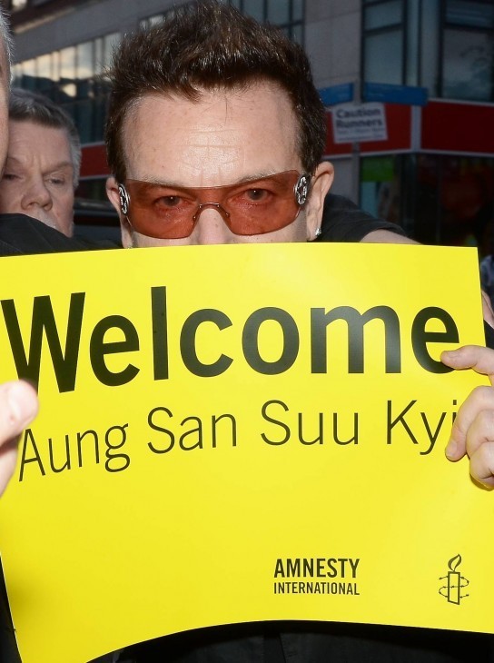 Photo of Bono, his face half-hidden behind a sign reading 'WELCOME Aung San Suu Kyi'