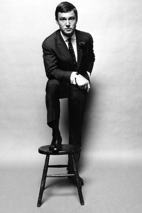 A photo of Vidal Sassoon, in skinny black suit, standing with one foot on a stool