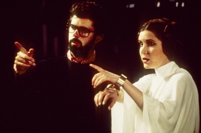 George Lucas and Carrie Fisher talk, both pointing off-camera, Carrie dressed as Leia
