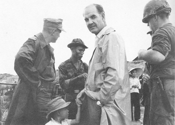 Photo of George McGovern in a trench coat, standing amid soldiers