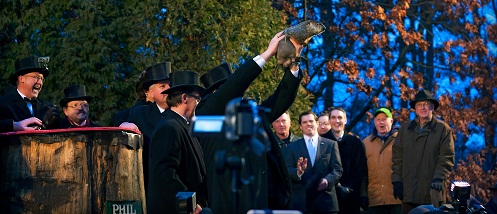 Four guys in top hats hold up an uncomfortable-looking groundhog as the crowd cheers
