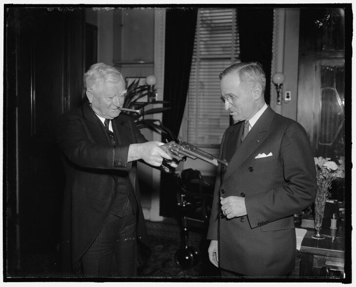 Photo of Harry Truman looking at two long pistols held by a grinning white-haired dude with a cigar